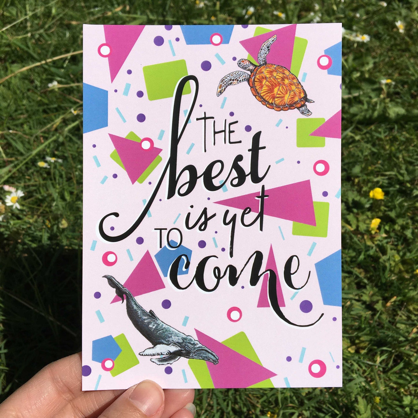 Photo of an A6 Postcard featuring a design (with a pale pink background covered in shapes: green squares, pink triangles, blue hexagons, pink and white dots, purple dots, and light blue confetti lines. A sea turtle is top right, a blue whale bottom left, and the words 'The best is yet to come' in the centre) from the Positivity Collection by Jenny Pond, JPArtwork