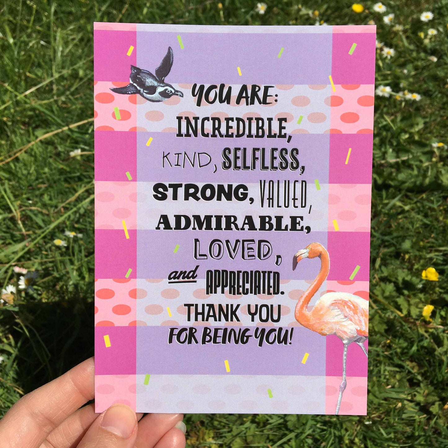 Photo of an A6 Postcard featuring a design (with a striped background of alternating pink stripes with pale pink and coral spotted stripes, yellow and green confetti lines fall down the page, and a light blue wide transparent strip down the centre backs the wording 'You are: incredible, kind, selfless, strong, valued, admirable, loved, and appreciated. Thank you for being you!' A penguin is top left, and a flamingo is bottom right) from the Positivity Collection by Jenny Pond, JPArtwork
