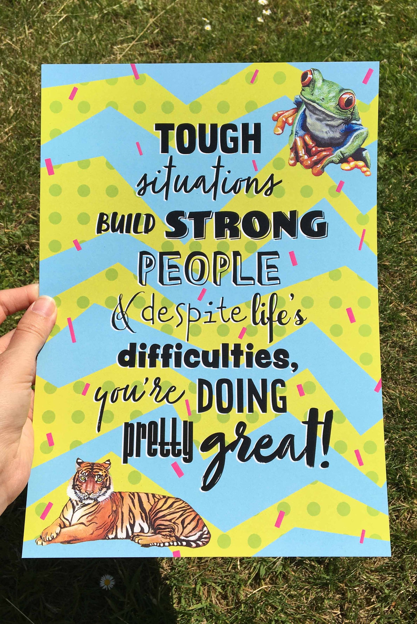 Photograph of an A4 Print; the design has a spotty green, and blue zigzag background covered in pink confetti lines, a tree frog top right, a tiger bottom left, and the words 'Tough situations build strong people & despite life's difficulties, you're doing pretty great!' in the centre. Artwork by Jenny Pond, JPArtwork
