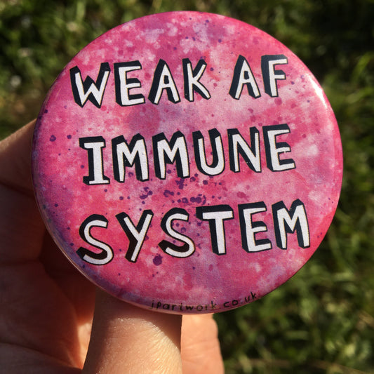 Photo shows a large pink pin badge with the words WEAK AF IMMUNE SYSTEM in bold 3D writing on. Ideal badge for the immunocompromised, and those with hidden illnesses/disabilities. By Jenny Pond, JP Artwork