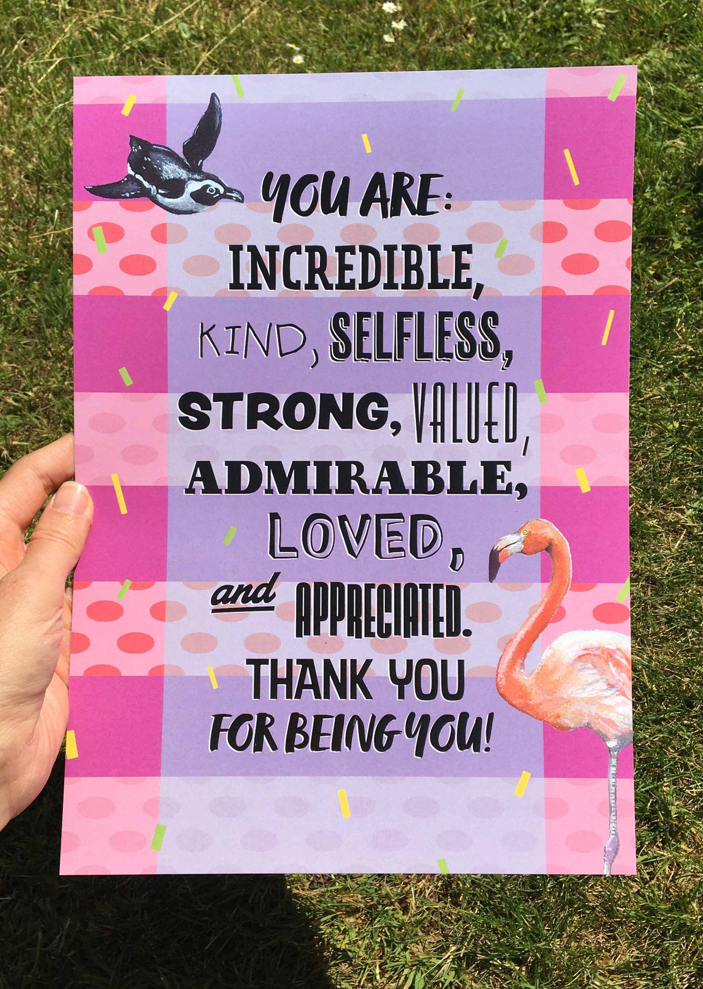 Photograph of an A4 Print featuring a design (with a striped background of alternating pink stripes with pale pink and coral spotted stripes, yellow and green confetti lines fall down the page, and a light blue wide transparent strip down the centre backs the wording 'You are: incredible, kind, selfless, strong, valued, admirable, loved, and appreciated. Thank you for being you!' A penguin is top left, and a flamingo is bottom right) from the Positivity Collection by Jenny Pond, JPArtwork